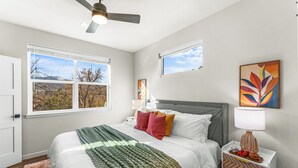 Un dormitorio con cama, mesita de noche, lámpara y ventana con vista a árboles y montañas.