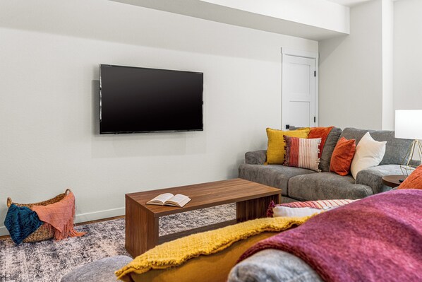 Una sala de estar moderna con un televisor de pantalla plana montado en la pared, un sofá gris con cojines coloridos, una mesa de centro de madera con un libro abierto y una alfombra estampada.
