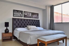 Een slaapkamer met een groot bed, een nachtkastje, een bankje en drie ingelijste foto’s aan de muur.