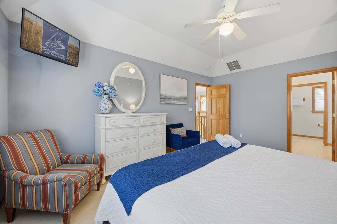 A bedroom with a bed, a dresser, a chair, a ceiling fan, and a door.