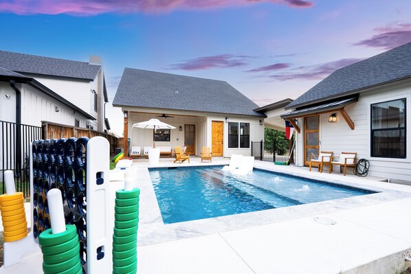 A residential swimming pool with a patio area, outdoor furniture, and a flag.