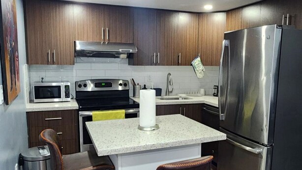 A modern kitchen with wooden cabinets, stainless steel appliances, a central island with stools, and a trash can.