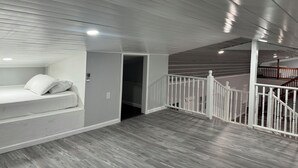 A spacious room with a wooden floor, a bed with white bedding, a white railing, and a staircase leading to an upper level. - Charming 4-bedroom barndominum in Elberta with AC, WiFi, 5 miles from boat ramp
 (Elberta)