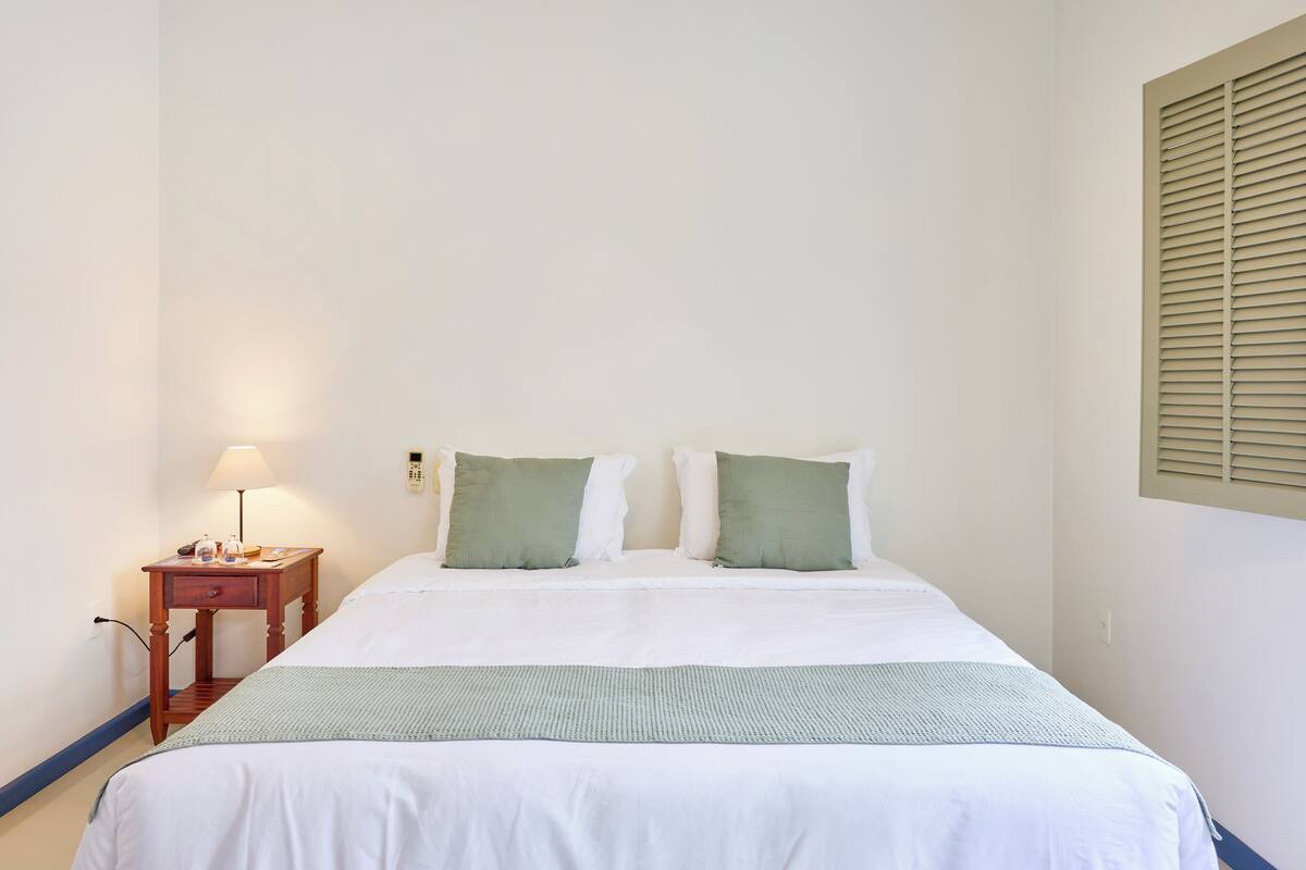 Un lit bien fait, avec une literie blanche et un plaid vert, une table de chevet avec une lampe, et un téléphone fixé au mur.