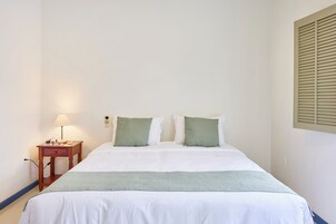 Een keurig opgemaakt bed met wit beddengoed en een groene plaid, een nachtkastje met een lamp, en een telefoon bevestigd aan de muur.