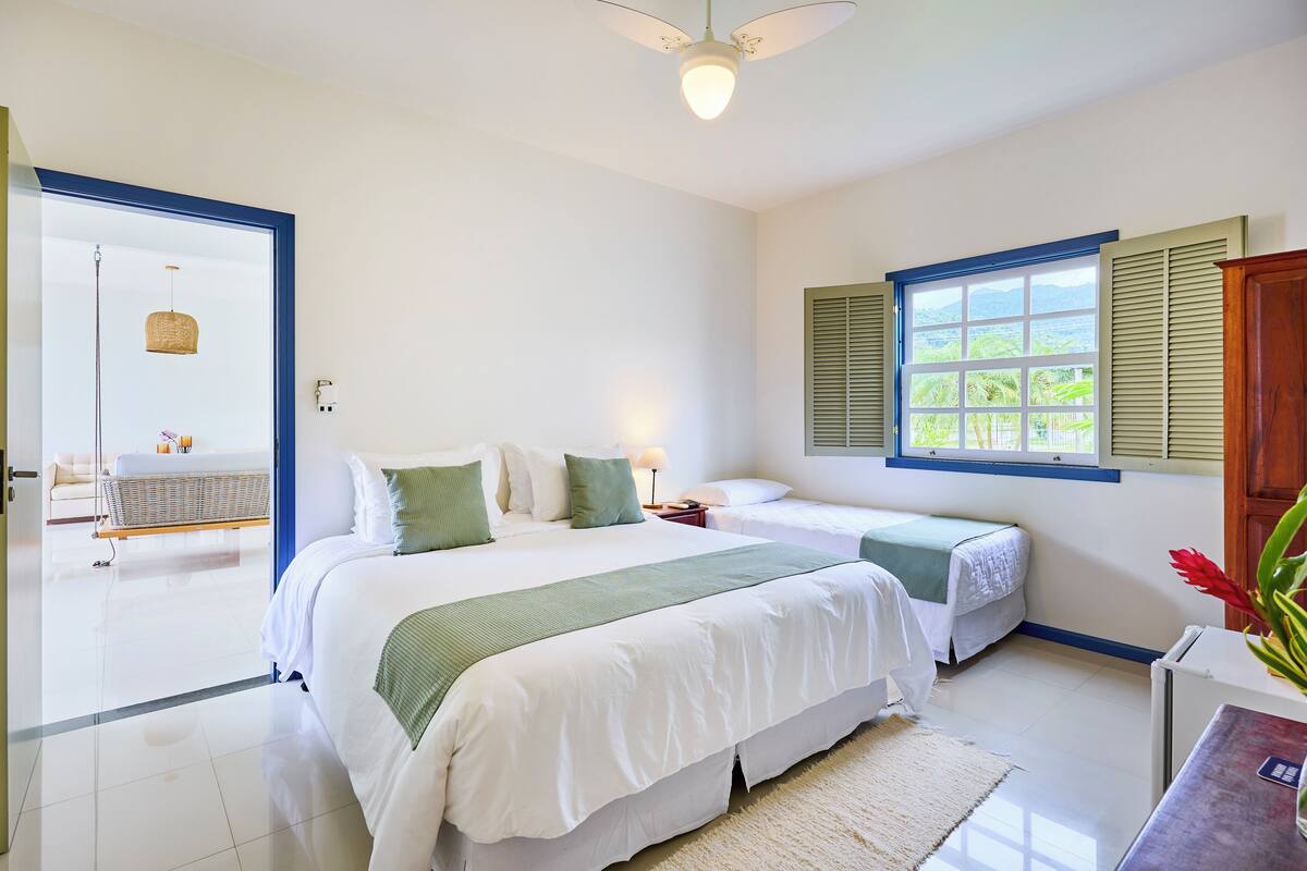 Une chambre d’hôtel avec deux lits, une grande fenêtre avec volets, un ventilateur de plafond et une vue sur l’extérieur.