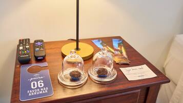 Una mesita de noche de madera con dos copas de vidrio que contienen grageas de chocolate, un control remoto, un mantel individual amarillo y una tarjeta con texto.