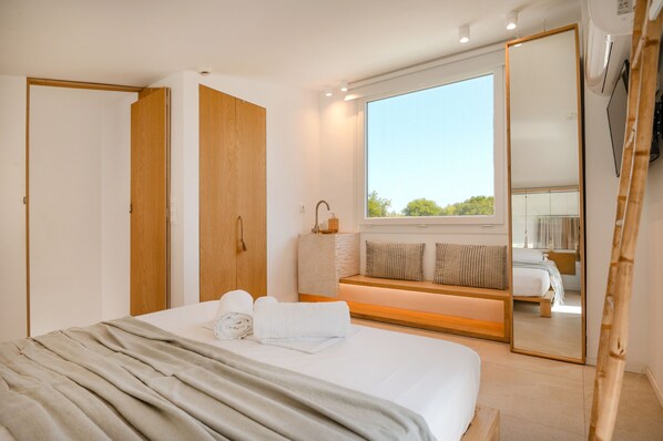 A modern bedroom with a large bed, a bench with pillows, a wooden wardrobe, and a large mirror.