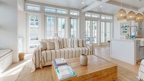 Una sala de estar amplia con un sofá grande, una mesa de centro de madera y una zona de cocina con encimera blanca y lámparas colgantes.