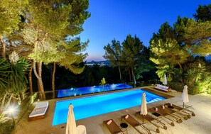 Piscina con sillas de descanso y sombrillas, rodeada de árboles y vista a la ciudad al atardecer.