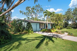 A single-story house with a light blue exterior, surrounded by lush greenery and palm trees. - ⚓Palma Sola Oasis!  Cozy tropical home w/ Putting Green & Hot tub minutes from AMI!⚓ (Bradenton)
