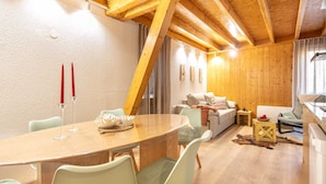 A cozy living room with a wooden ceiling, a dining table with chairs, a sofa, and a small side table with a candle.