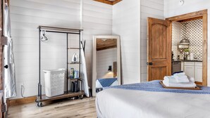 Un dormitorio con cama, una puerta de madera, un carro de lavandería y una pequeña cocina americana.