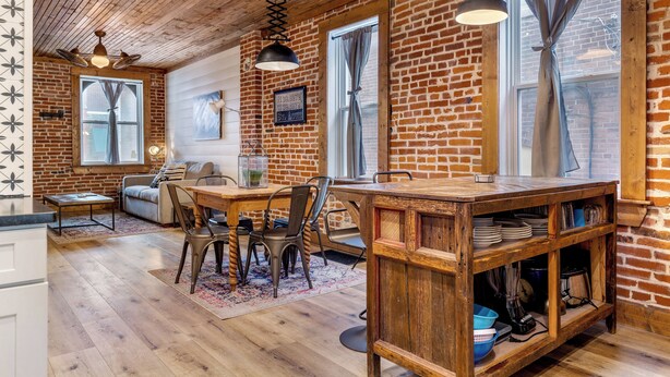 Una cocina rústica con vigas de madera, un comedor con mesa y sillas, y una pared de ladrillo.
