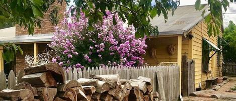 Una casa de madera rústica con un muro de piedra, un arbusto de flores rosadas y una valla de madera.