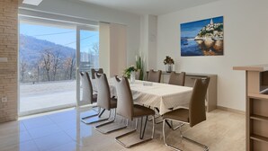 Un coin repas avec une table dressée pour six personnes, une grande fenêtre donnant sur des arbres et des collines, et un tableau accroché au mur.