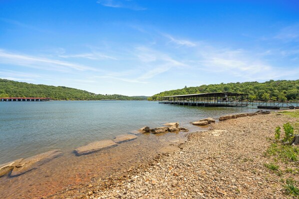 A lakeside view with a dock extending into the water, surrounded by rocky terrain and lush greenery. - Charming Escape with Scenic Wood Views, Walk-to-Lake Access, Outdoor Pool & Free Attraction Tickets (Kimberling City)