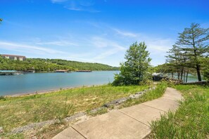 A concrete pathway leading to a body of water with a forested hill in the background. - Charming Escape with Scenic Wood Views, Walk-to-Lake Access, Outdoor Pool & Free Attraction Tickets (Kimberling City)