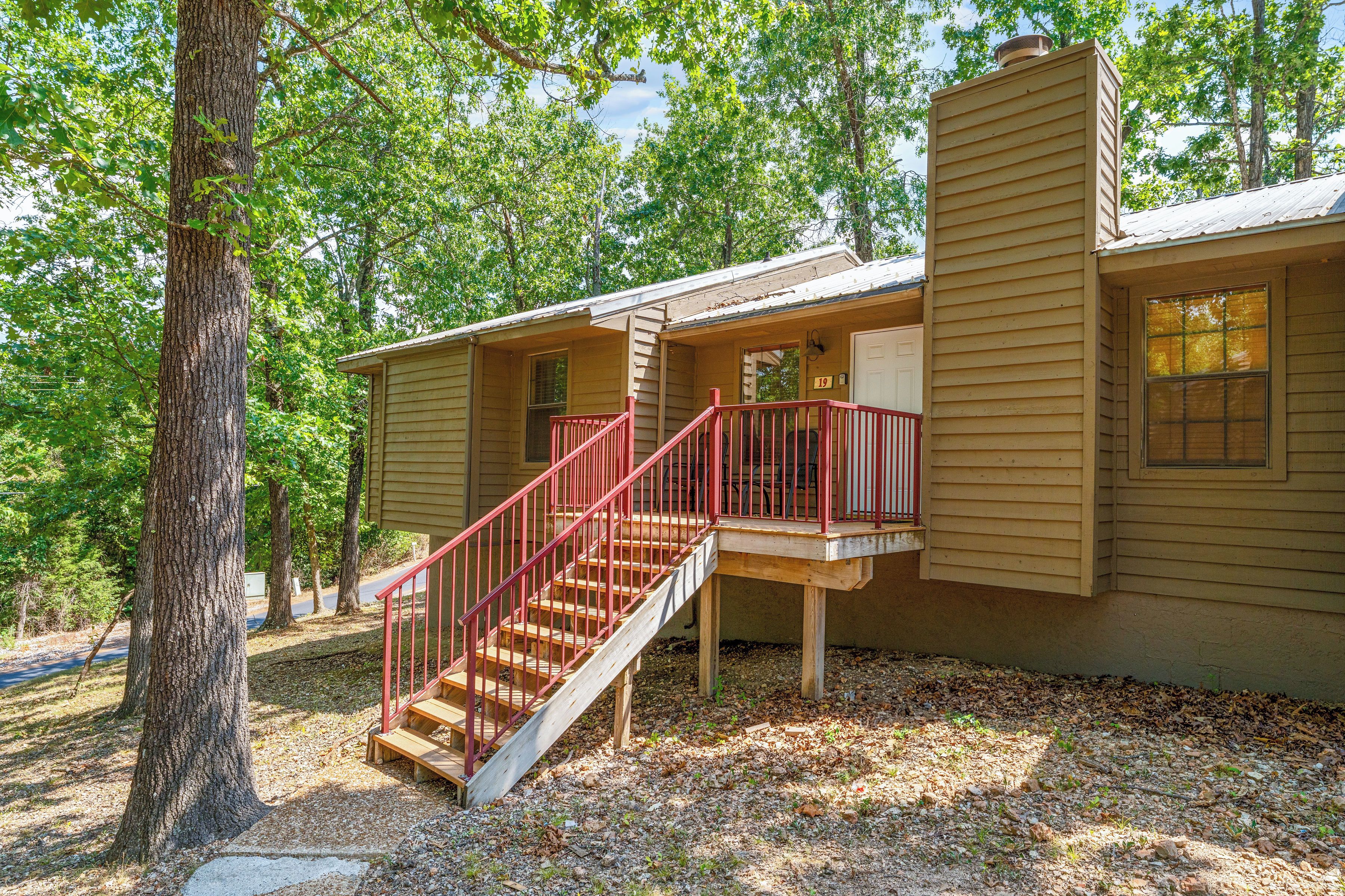 Une maison à deux étages avec un escalier en bois et une rampe rouge, entourée d’arbres.