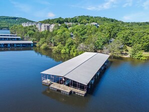 A floating structure with a covered deck, surrounded by calm water, with buildings and trees in the background.