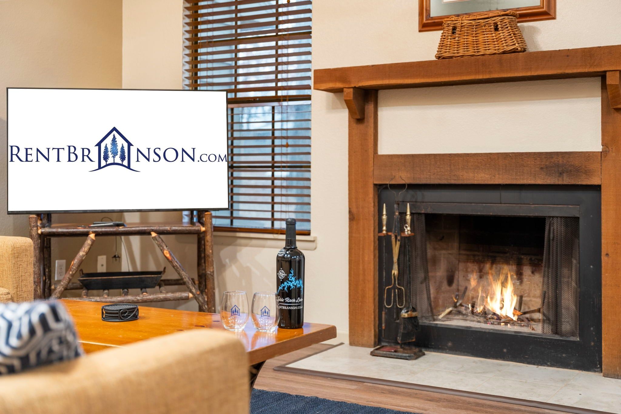 A cozy living room with a fireplace, a table with a bottle and glasses, and a logo for RENTBRINSON.COM.