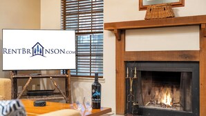 Una sala de estar acogedora con chimenea, una mesa de madera con vasos y una botella, y un cartel de RENTBRINSON.COM.