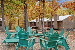 Un camping con sillones Adirondack verdes dispuestos alrededor de un hogar central, rodeado de árboles con follaje otoñal.