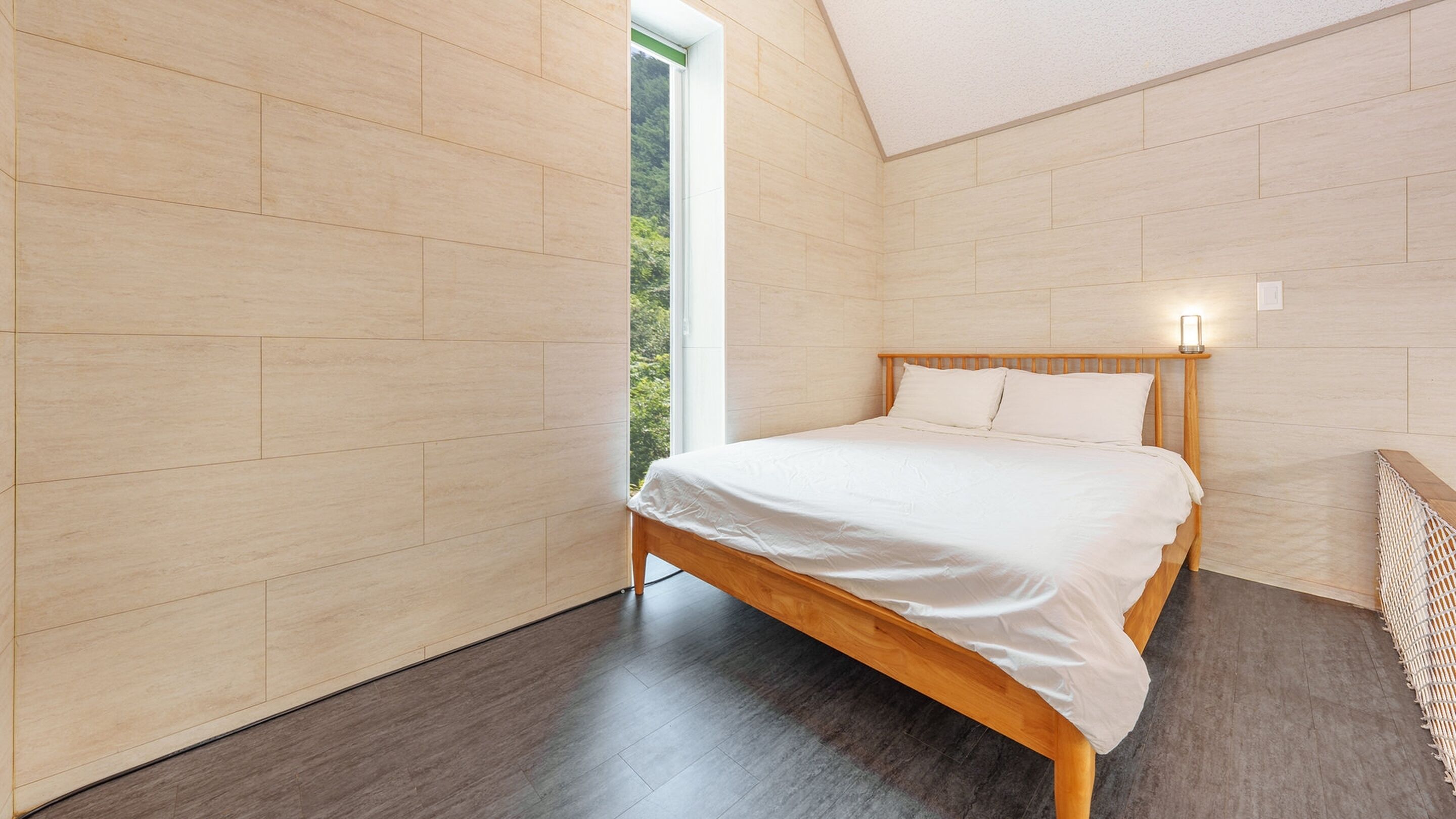 A modern bedroom with a wooden bed, a view of greenery through a window, and a sloped ceiling.