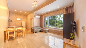 A modern living room with a sofa, a dining table with chairs, a TV, and a large window with a view of trees.