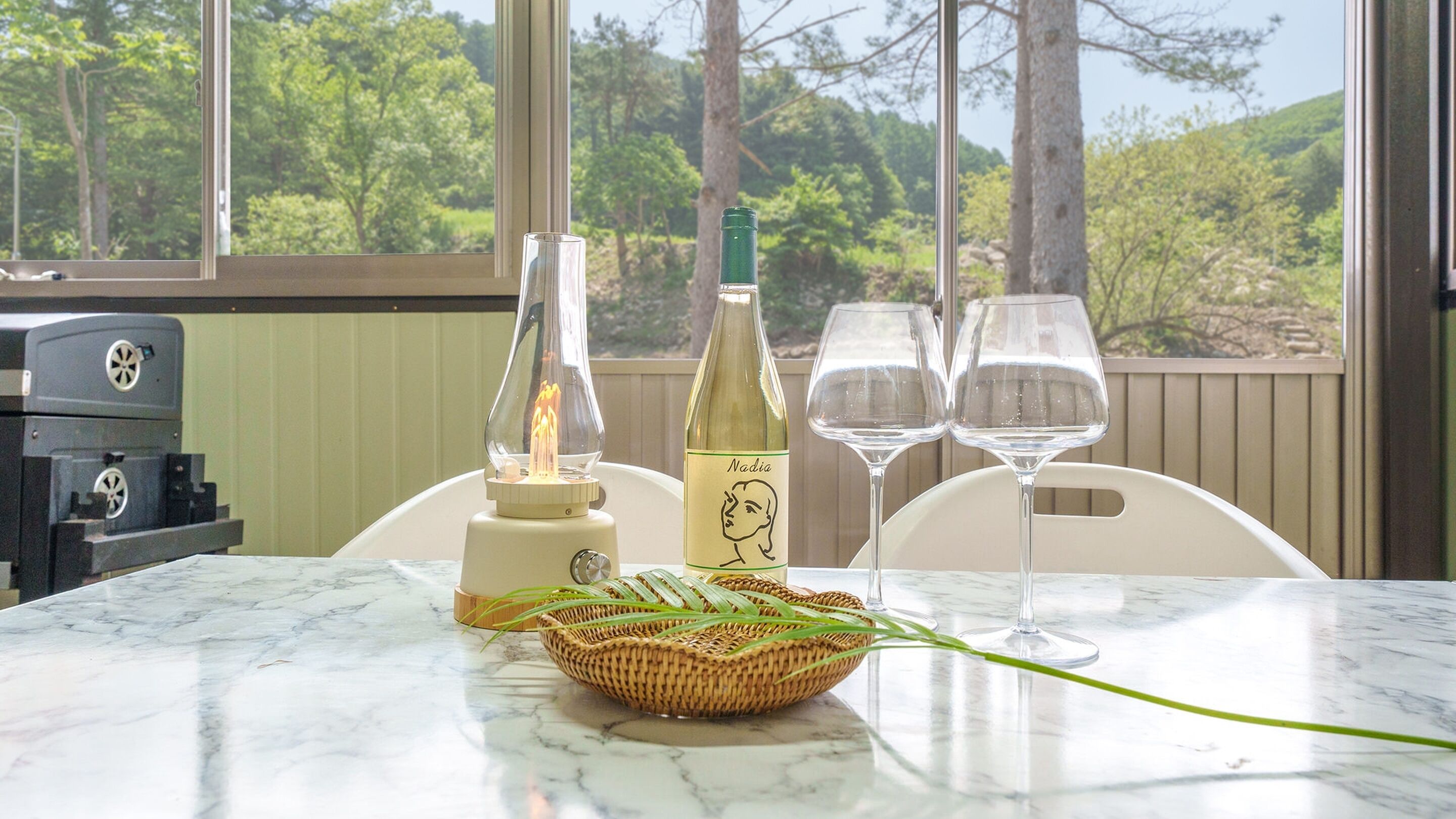 A table with a bottle of wine, two glasses, a candle, and a basket of green leaves.