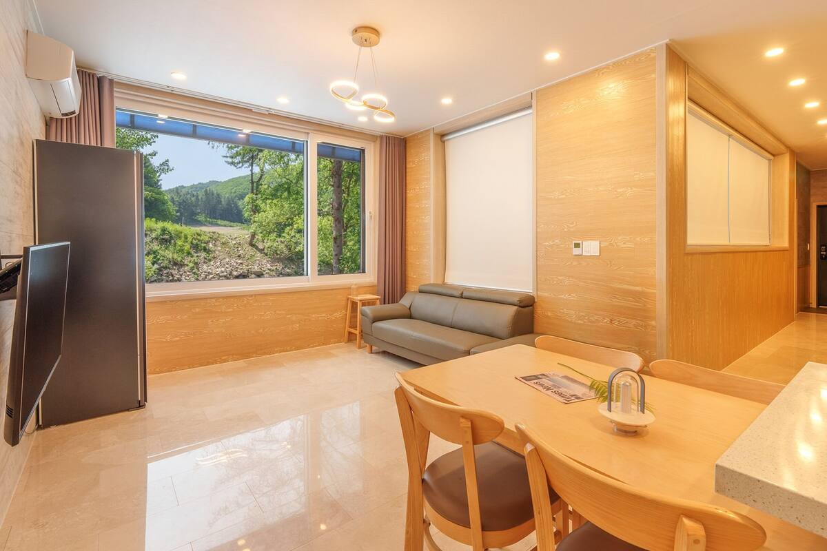 Eine moderne Küche mit einem eingebauten Kühlschrank, einem Esstisch mit Stühlen, einer Couch und einem großen Fenster mit Blick ins Grüne.