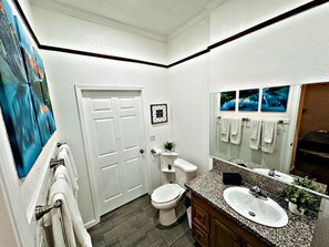 Een badkamer met een wit toilet, een wastafel en een spiegel. Handdoeken hangen aan een rek en er hangt een fotolijst aan de muur.