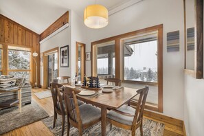 Comedor con mesa y sillas de madera, ventanas amplias con vista a árboles y una lámpara colgante.
