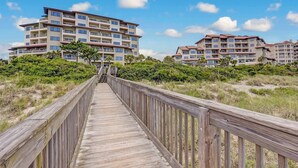 A wooden boardwalk leading to a multi-story hotel with balconies, surrounded by greenery and under a clear sky.