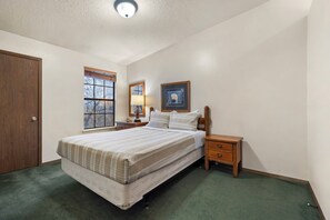 Ein Schlafzimmer mit einem Bett, einem Nachttisch, einer Lampe, einem gerahmten Bild und einem Fenster.