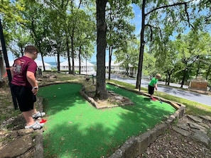 Un campo de mini golf con superficie de juego verde, rodeado de árboles y un edificio al fondo.