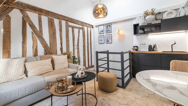 En moderne stue med sofa, sofabord og skammel, et køkkenområde med spisebord, samt en hængende lampe.
