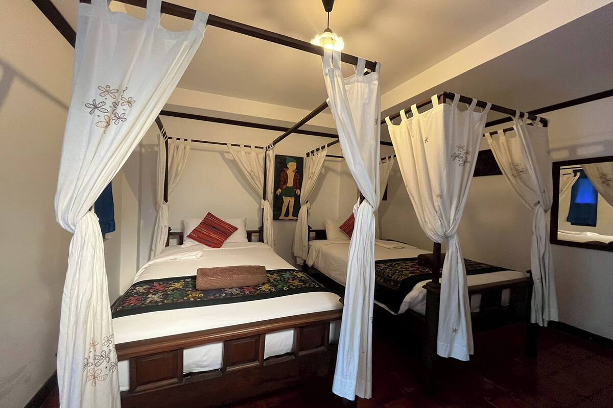 A room with two beds, each with a canopy bed frame and white curtains. There is a painting on the wall and a window with a view of the outside.