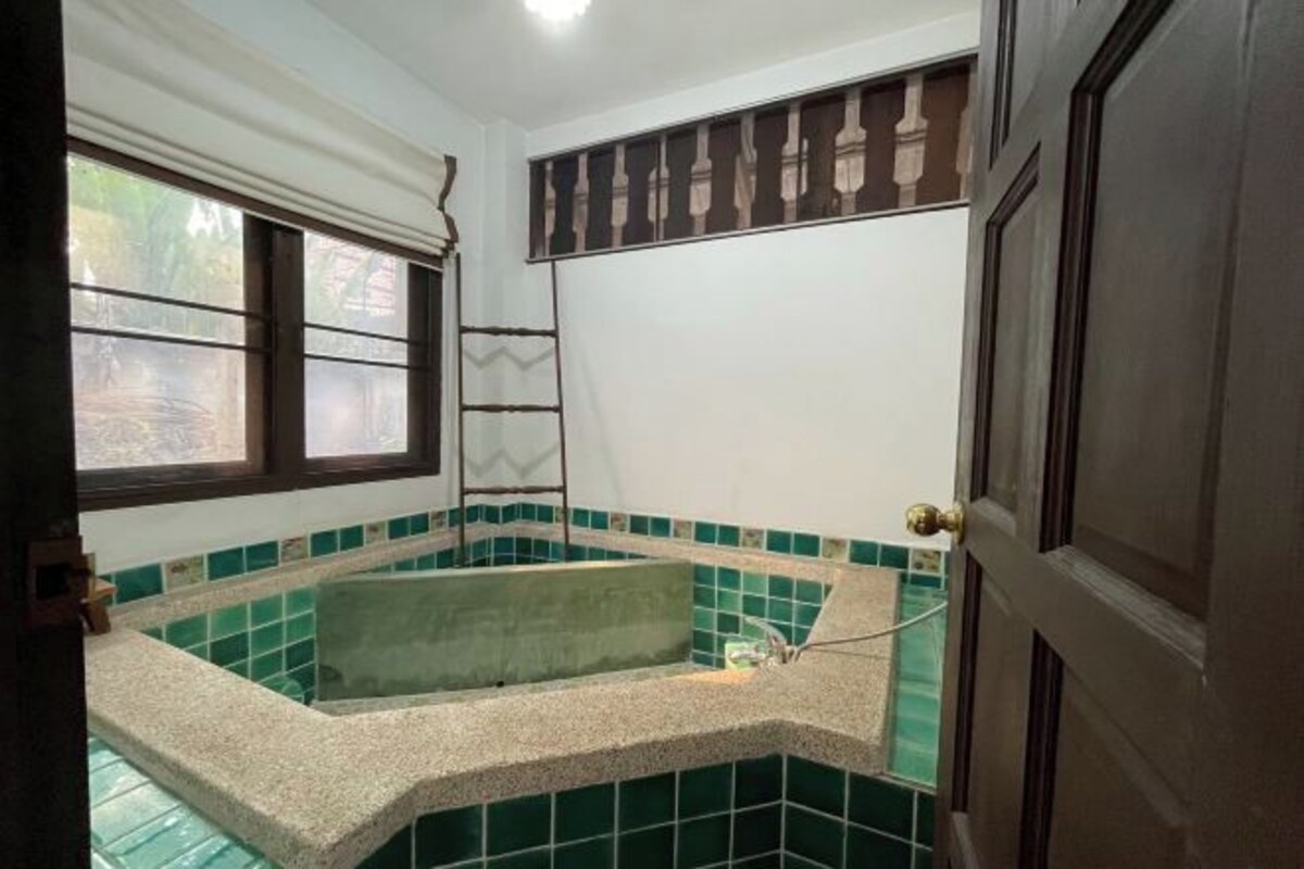 Une salle de bain avec une baignoire d’angle, des murs recouverts de carreaux verts, une fenêtre avec des stores et une porte dotée d’une poignée en laiton.