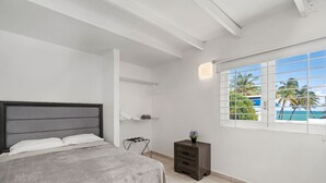 Un dormitorio con cama, mesita de noche, ventana y vista a palmeras y cielo azul.