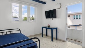 Una habitación con cama, una mesita con un florero, un televisor en la pared y una ventana con vistas a edificios.