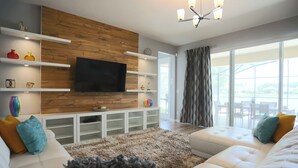 A modern living room with a flat-screen TV mounted on a wooden wall, white sofas, a fluffy rug, and a large window with curtains. - Pool Villa, Game Room, Theater, 20 Minutes to Disney, Great Pool Heat (Kissimmee)
