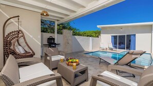 An outdoor patio with wicker furniture, a hanging chair, a pool, and a dining table with fruit.