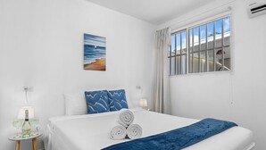 A hotel room with a large bed, a window with curtains, a painting of a beach scene, and a small table with a lamp and glasses.