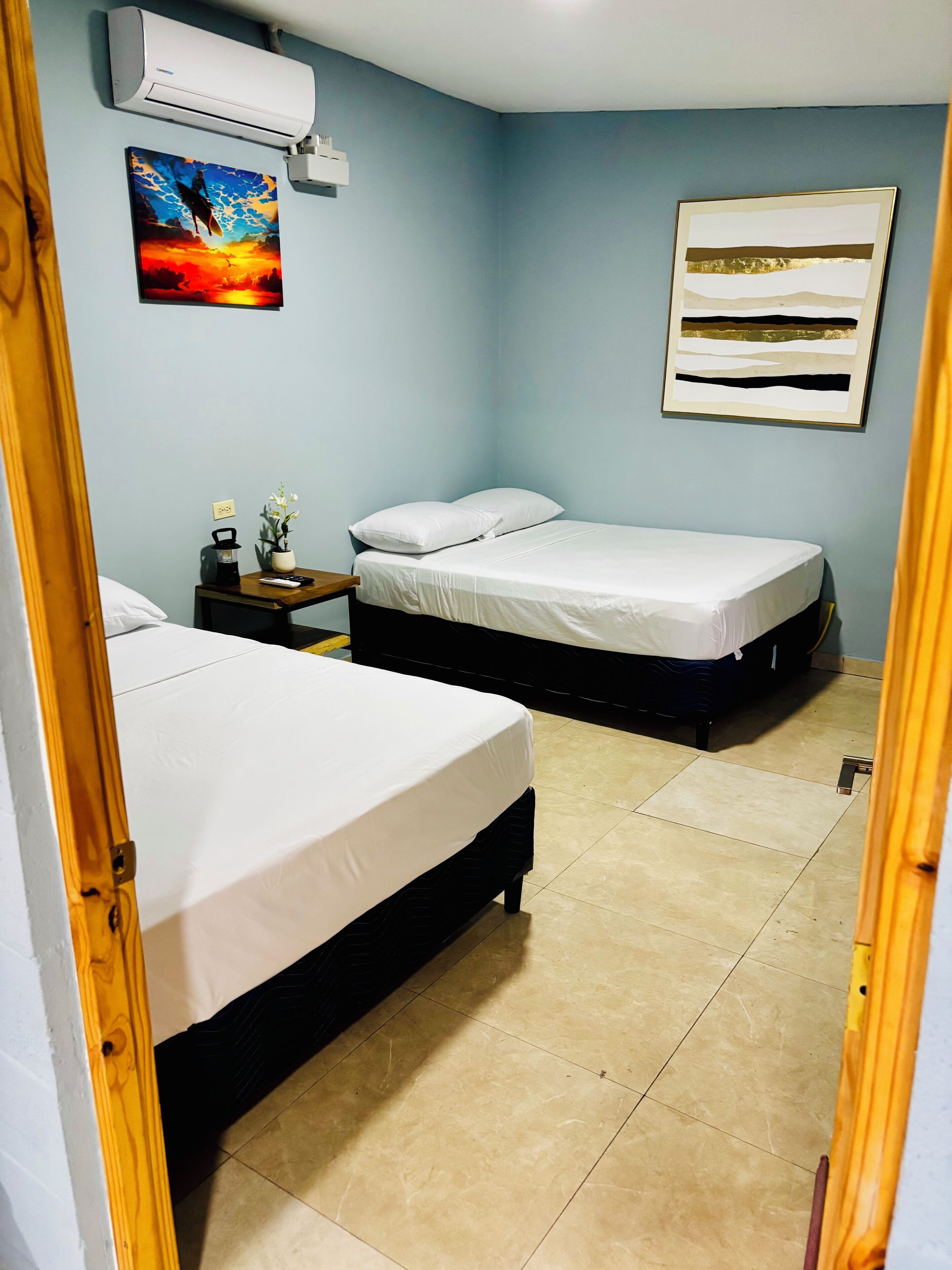 A bedroom with two beds, a nightstand, a wall-mounted air conditioner, and a painting.
