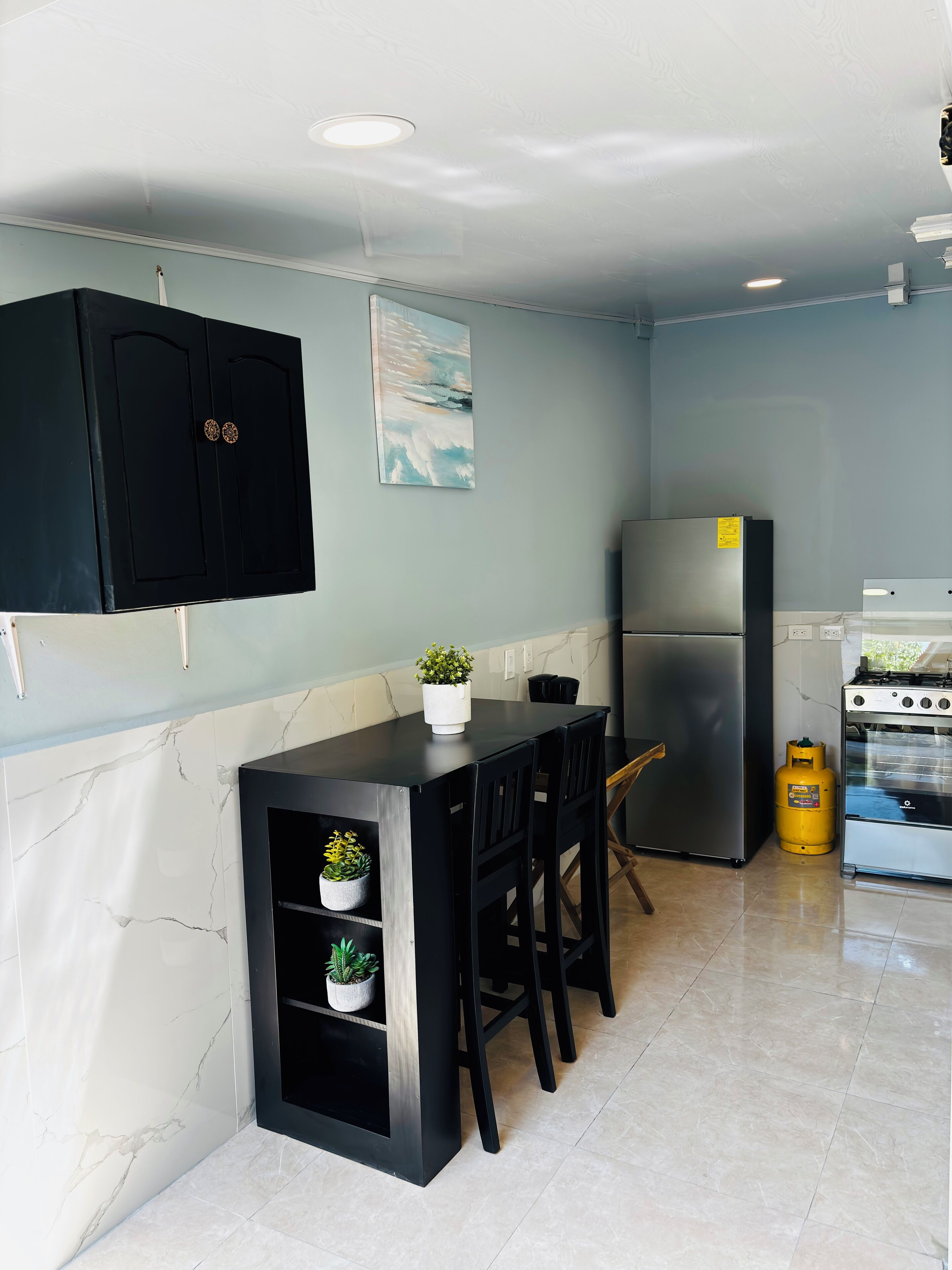 Una cocina moderna con salpicadero de mármol, armarios negros, un refrigerador de acero inoxidable, una estufa a gas y un pequeño comedor con sillas y plantas en macetas.