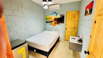 A bedroom with a bed, a wooden door, a wall-mounted air conditioner, and a small table with a remote control and flowers.