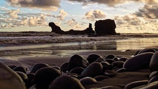 A beach with large boulders and a rocky outcrop in the distance under a partly cloudy sky.