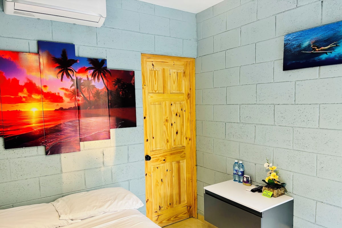 A room with a bed, a wooden door, a small table with a water bottle and a flower, and three sunset paintings on the wall.