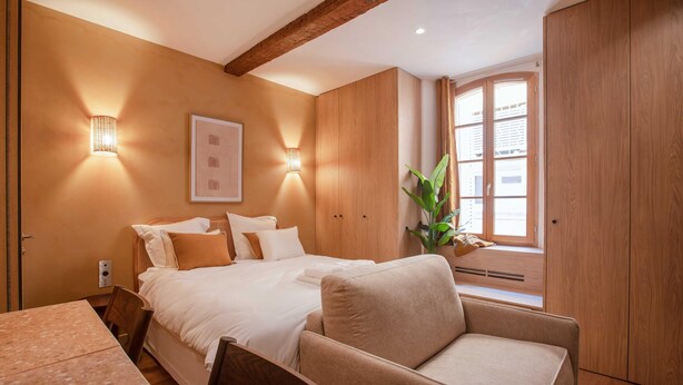 A bedroom with a bed, a chair, a table, a wall-mounted light, and a window with curtains. - Charming Studio With A/C - 2P - Center St Tropez (Saint-Tropez)
